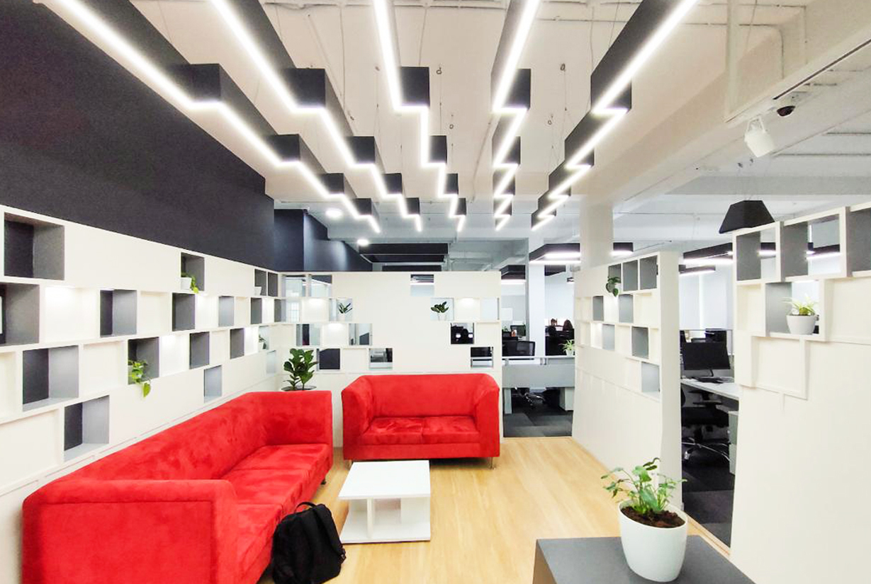 Office lounge area with vibrant red sofas, modern ceiling lights, and
						 decorative wall shelving