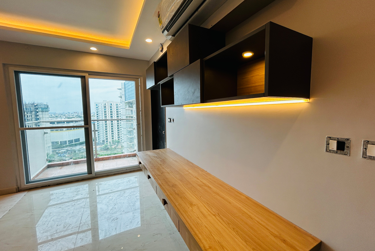 Bright workspace with a wooden desk, open shelving,
						 ambient lighting, and a balcony view overlooking high-rise buildings.